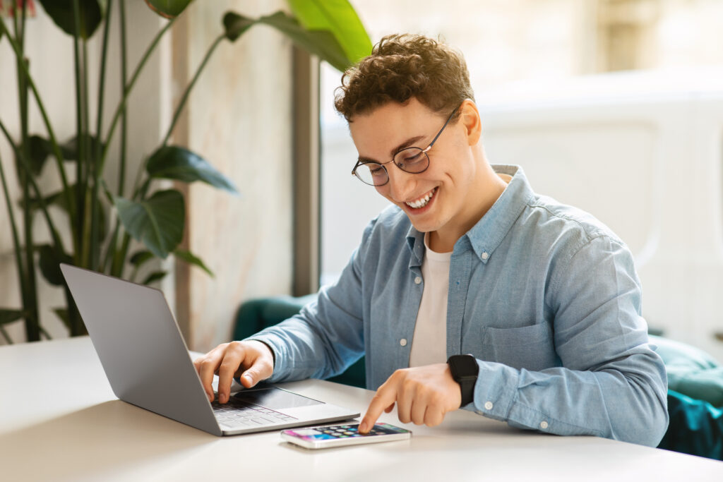 Happy young european guy student in glasses, typing on smartphone, laptop, work, study online, chatting in cafe. Online meeting app, modern lifestyle remotely, digital nomad